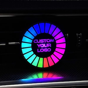 Puede incluir: Un accesorio circular para la ventilación del coche con un anillo de luz LED multicolor vibrante que cambia de azul a verde y rosa. El centro del dispositivo es negro y muestra el texto iluminado en violeta CUSTOM YOUR LOGO.