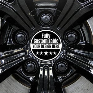 Puede incluir: Primer plano de una rueda de coche negra con un tapacubos personalizado. El tapacubos es blanco con el texto "Fully Customizable YOUR DESIGN HERE" y cinco estrellas. La rueda tiene un acabado brillante.