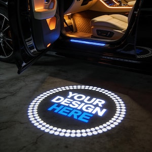 May include: A car door is open, revealing interior lighting and a custom door projector. The projector displays "YOUR DESIGN HERE" in white and blue on the ground. The car's interior features beige seating and ambient lighting.