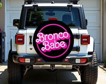 May include: A white SUV with a black spare tire cover. The cover has the words "Bronco Babe" in pink script inside a pink circle on a black background. The vehicle has a roof rack and is parked in front of a garage.