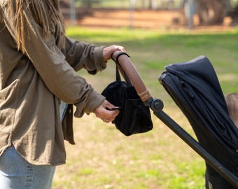 May include: A black stroller with a brown handle and a black accessory bag. A person is holding the bag. A child is seated in the stroller. The background is a grassy area.