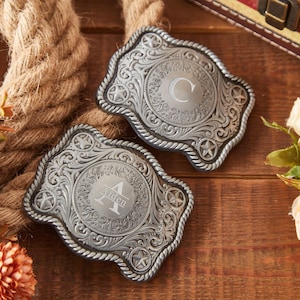 May include: Two silver belt buckles with ornate designs and engraved initials. The buckle on the left has the initial "A" and the word "Aiden" engraved on it. The buckle on the right has the initial "C" engraved on it.