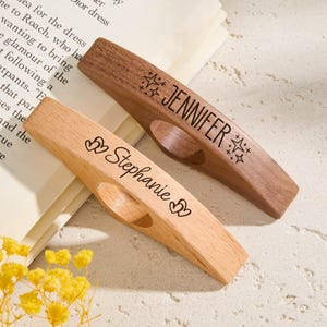 May include: Two wooden book page holders, one light-colored and one dark-colored. The light-colored holder has the name "Stephanie" engraved, and the dark-colored holder has "Jennifer". Both have a star design.