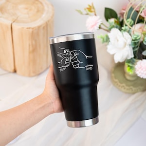 May include: Black stainless steel tumbler with a silver rim and base. The tumbler features a white graphic of three fists bumping, with the names "Milo", "Josh", and "Dad" inscribed. The tumbler is designed to hold approximately 887 ml.