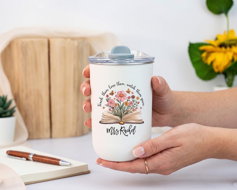 Pu&ograve; includere: Bicchiere termico bianco con coperchio grigio, tenuto in mano da una persona. Il bicchiere presenta un motivo floreale con un libro aperto e il testo "Teach them, love them, watch them grow" e "Mrs. Rachel". Una penna e un taccuino sono sullo sfondo.