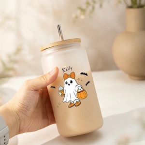 Puede incluir: Un vaso de vidrio esmerilado con tapa de bambú y pajita de metal. El vaso presenta un gráfico de un fantasma blanco con lazo y zapatos naranjas, sosteniendo un café y un cubo de calabaza, junto al nombre Kelly y pequeños murciélagos negros.