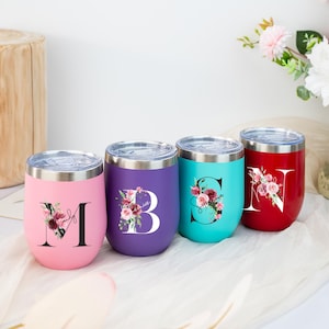 Könnte beinhalten: Vier isolierte Weingläser in Rosa, Lila, Türkis und Rot. Jeder Becher hat einen großen Anfangsbuchstaben mit floralem Design. Die Becher haben einen silbernen Rand und einen transparenten Deckel. Der rosa Becher hat den Buchstaben "M" und den Namen "Mandy".