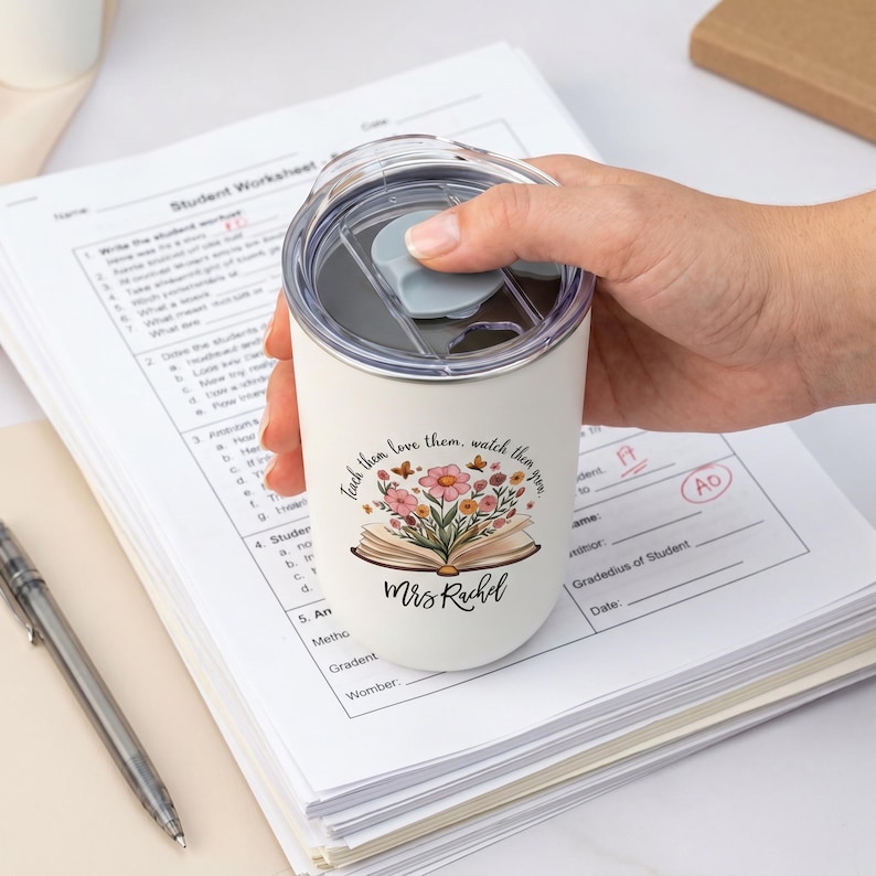 Pu&ograve; includere: Un bicchiere termico bianco con coperchio nero, tenuto in mano. Il bicchiere presenta la scritta "Teach them, love them, watch them grow" con un design floreale e di libro, e il nome "Mrs Rachel".