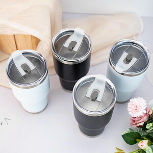 May include: Four stainless steel tumblers with a tapered design, featuring two matte black and two light blue finishes. Each tumbler includes a clear, removable lid with a white sliding closure mechanism for the drinking opening.