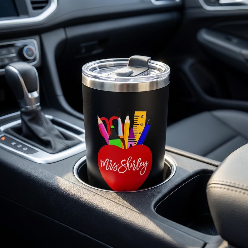 Pu&ograve; includere: Un bicchiere da viaggio nero con bordo e coperchio argentati, con un design a mela rossa con materiale scolastico e il nome "Mrs. Shirley" in corsivo bianco. Il bicchiere &egrave; nel portabicchieri di un'auto.