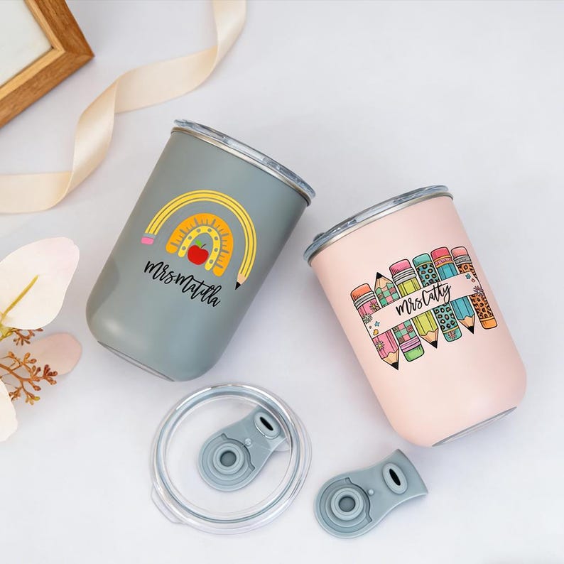 Pu&ograve; includere: Due bicchieri isolati con coperchi. Uno &egrave; grigio con un design arcobaleno e mela, il testo "Mrs. Matilda". L'altro &egrave; rosa con grafica di matite e il testo "Mrs. Cathy".