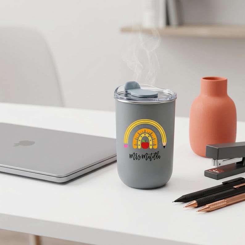 Pu&ograve; includere: Una tazza da viaggio grigia con un disegno di arcobaleno, mela e matita. Il testo "Mrs. Matilla" &egrave; sotto. La tazza ha un coperchio grigio e si trova su una superficie bianca con un laptop, penne e una pinzatrice.