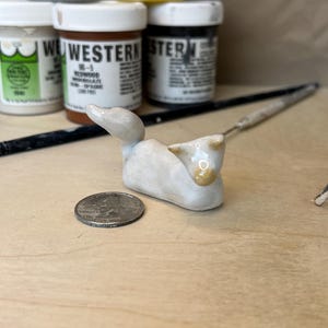 May include: A small, white ceramic duck figurine with gold accents, next to a U.S. quarter. Paint jars in the background include colors like silver gray, bright yellow, and redwood.