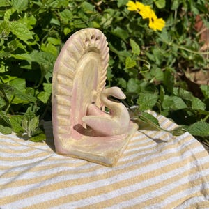 May include: A ceramic bookend featuring a sculpted swan figure centered in front of a semi-circular, ribbed back panel. The piece is finished in a glossy, pale pink and cream glaze with subtle variations in color across the textured surface.