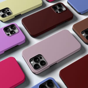 Puede incluir: Una colección de fundas de teléfono coloridas en varios tonos, incluyendo rosa, morado, amarillo, marrón y azul. Cada funda tiene un acabado mate suave y un recorte para la cámara. Las fundas están dispuestas sobre un fondo claro.