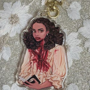 Op de afbeelding: Een heldere acryl sleutelhanger met een gestileerde illustratie van een vrouw met donker haar en een crèmekleurige jurk, bespat met rood. Ze houdt een boek vast. De sleutelhanger heeft een gouden stervormige sluiting.