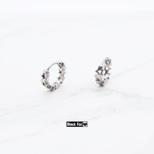 May include: Two silver hoop earrings, each adorned with star-shaped cutouts. The earrings are displayed on a white surface. The brand name "Black Fox" is printed in a black rectangle.