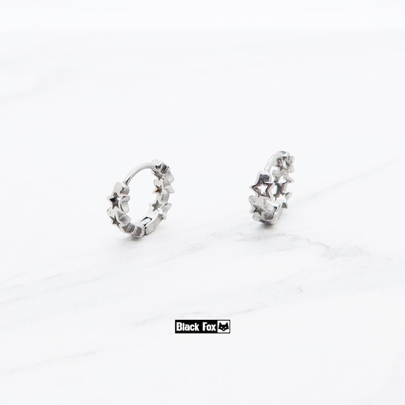 May include: Two silver hoop earrings, each adorned with star-shaped cutouts. The earrings are displayed on a white surface. The brand name "Black Fox" is printed in a black rectangle.