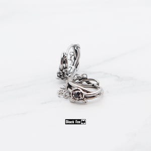 May include: Two silver hoop earrings with floral and vine detailing. Each earring has a small black stone at the centre of the flower design. The earrings are on a white background. The brand name "Black Fox" is in a black box.