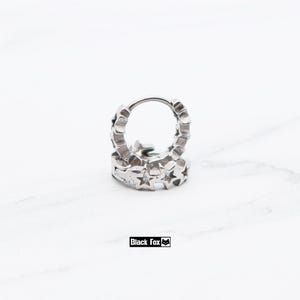 May include: A pair of silver-colored hoop earrings. Each earring is adorned with a series of star-shaped cutouts around the hoop. The earrings are displayed on a white surface. The brand name "Black Fox" is visible.