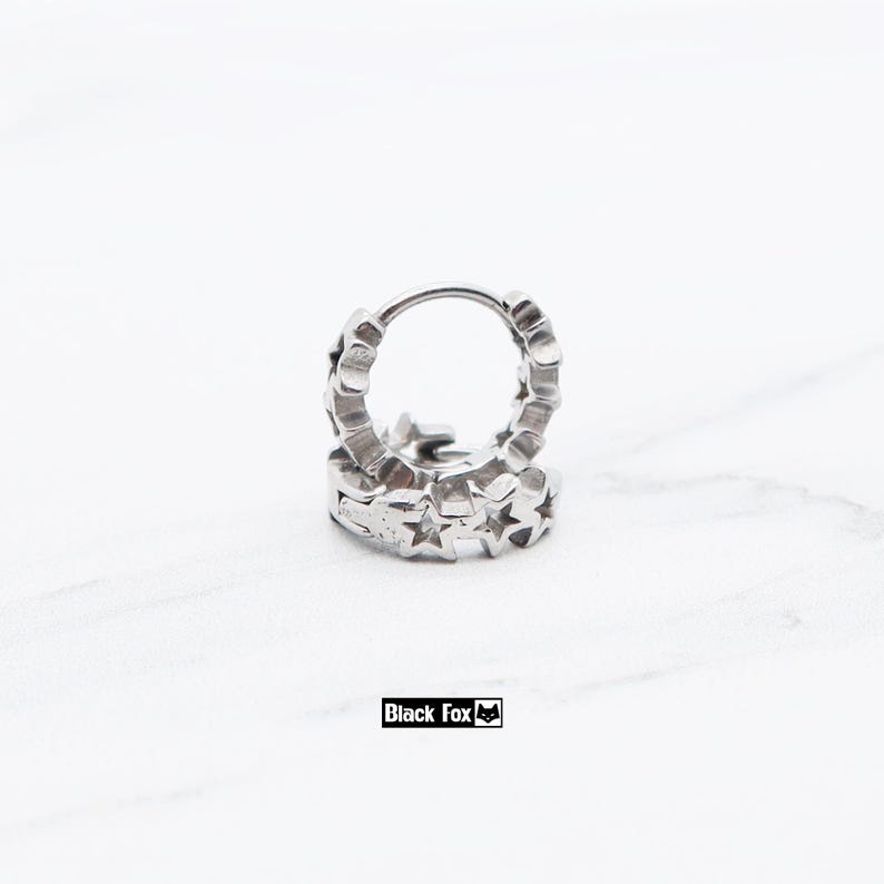 May include: A pair of silver-colored hoop earrings. Each earring is adorned with a series of star-shaped cutouts around the hoop. The earrings are displayed on a white surface. The brand name "Black Fox" is visible.