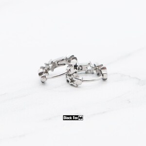 May include: A pair of silver hoop earrings. Each earring is adorned with star-shaped cutouts. The earrings are displayed on a white surface. The brand name "Black Fox" is printed in a black rectangle.