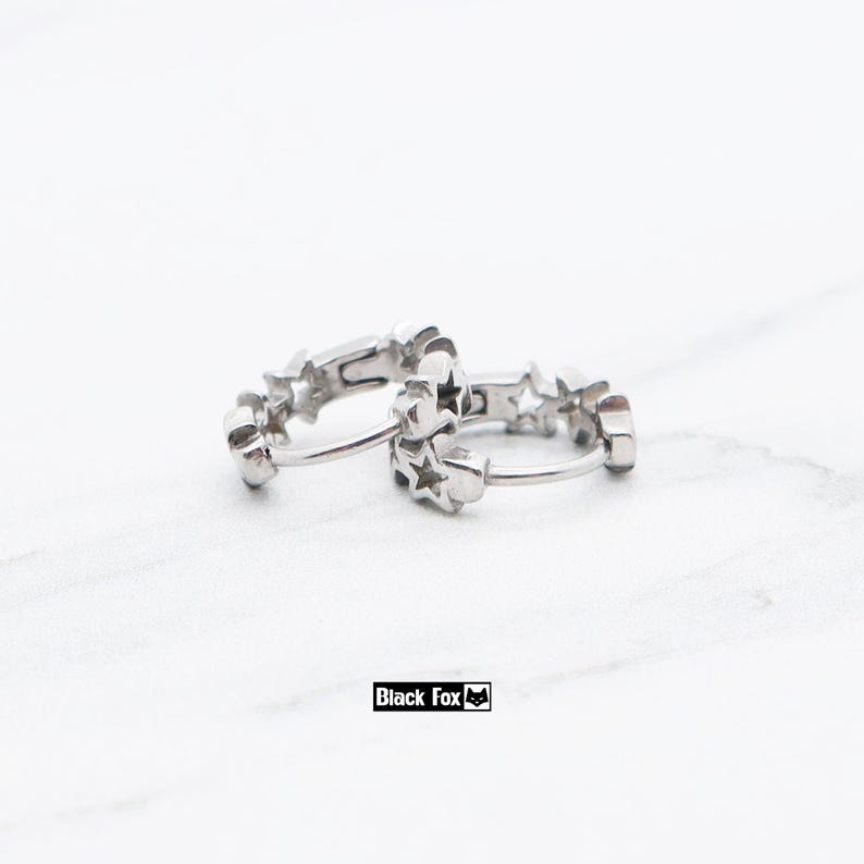 May include: A pair of silver hoop earrings. Each earring is adorned with star-shaped cutouts. The earrings are displayed on a white surface. The brand name "Black Fox" is printed in a black rectangle.