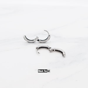 May include: Silver hoop earrings with a line of small, sparkling stones. The earrings are open, showing the clasp mechanism. The brand name "Black Fox" is printed in a black rectangle.