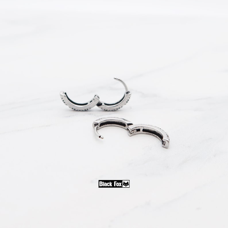 May include: Silver hoop earrings with a line of small, sparkling stones. The earrings are open, showing the clasp mechanism. The brand name "Black Fox" is printed in a black rectangle.