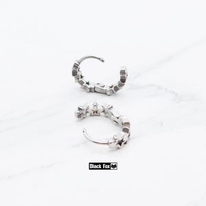 May include: Two silver hoop earrings with a star design. Each earring is a small, curved band with multiple star-shaped cutouts. The earrings are displayed on a white marble surface. The brand name "Black Fox" is printed in a black rectangle.