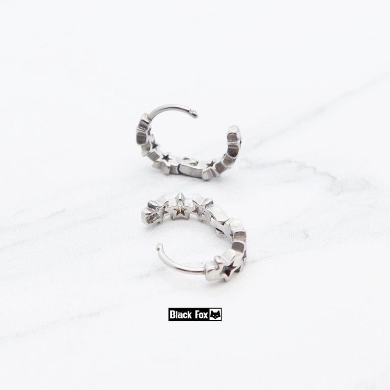 May include: Two silver hoop earrings with a star design. Each earring is a small, curved band with multiple star-shaped cutouts. The earrings are displayed on a white marble surface. The brand name "Black Fox" is printed in a black rectangle.