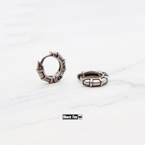 Könnte beinhalten: Zwei silberfarbene Creolen mit einem strukturierten, geometrischen Design. Die Ohrringe bestehen aus Metall und haben einen Klappverschluss. Die Ohrringe werden auf einer weißen Marmoroberfläche präsentiert. Der Markenname "Black Fox" ist sichtbar.