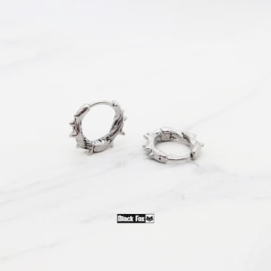 May include: Two silver hoop earrings with a barbed wire design, set against a white marble backdrop. The brand name "Black Fox" is displayed in a black rectangle. The earrings are a stylish accessory.