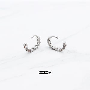 May include: A pair of silver hoop earrings with a star design. Each earring has a curved shape with small star cutouts along the outer edge. The earrings are displayed on a white surface. The brand name "Black Fox" is printed in a black rectangle.
