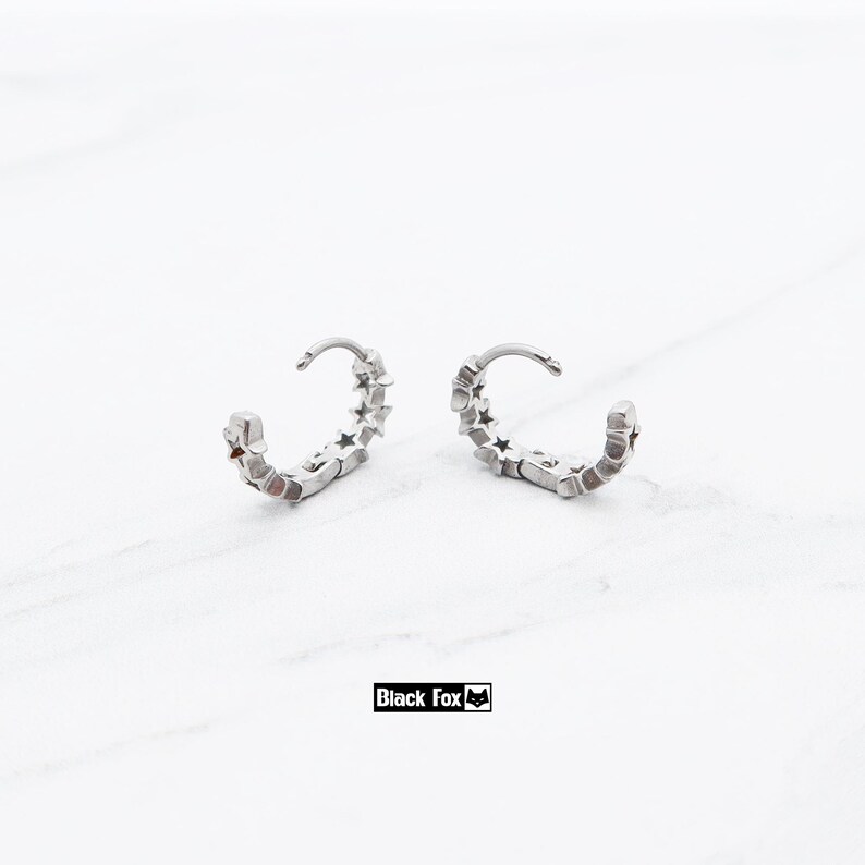 May include: A pair of silver hoop earrings with a star design. Each earring has a curved shape with small star cutouts along the outer edge. The earrings are displayed on a white surface. The brand name "Black Fox" is printed in a black rectangle.