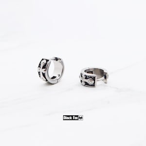 Peut inclure: Deux boucles d'oreilles créoles argentées avec un motif de croix décoratif. Les boucles d'oreilles sont en métal brillant et ont une fermeture à charnière. La marque "Black Fox" est imprimée dans un rectangle noir.