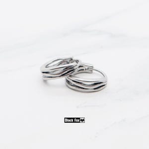 May include: A pair of silver hoop earrings with a layered, wavy design. The earrings are made of a shiny metal and are displayed on a white marble surface. The brand name "Black Fox" is printed in a black rectangle.