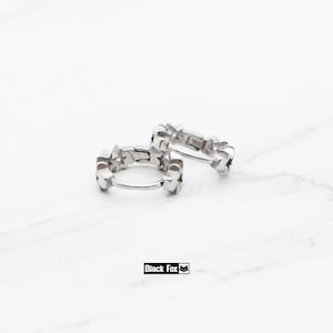 May include: Two silver hoop earrings with a star design. The earrings are made of a shiny metal and feature a series of small stars around the hoop. The earrings are displayed on a white surface. The brand name "Black Fox" is visible.