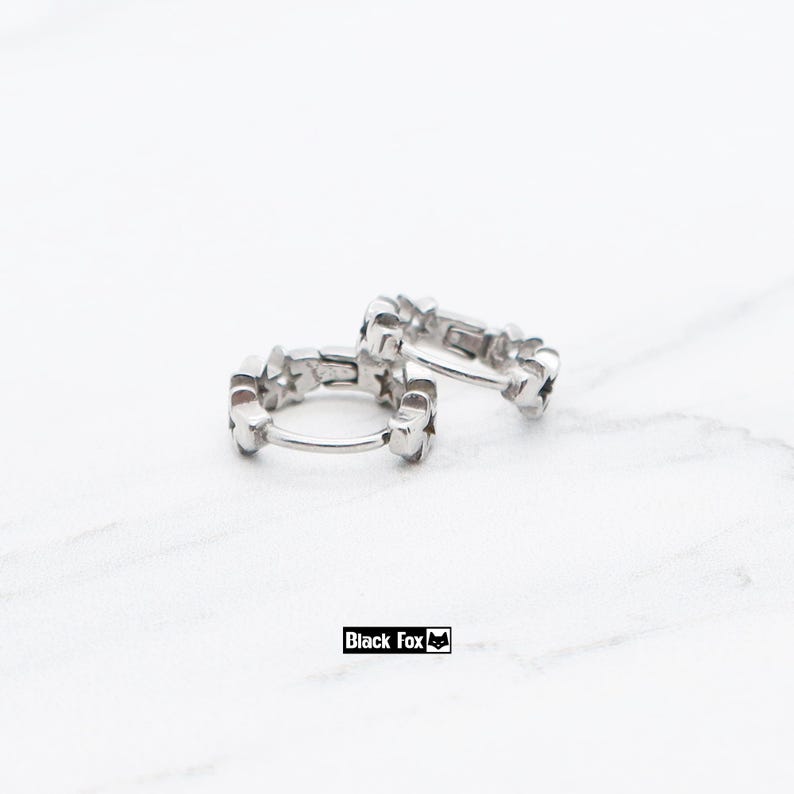 May include: Two silver hoop earrings with a star design. The earrings are made of a shiny metal and feature a series of small stars around the hoop. The earrings are displayed on a white surface. The brand name "Black Fox" is visible.