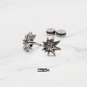 May include: A pair of silver-toned metal stud earrings shaped like seven-pointed leaves with detailed etched veins. The earrings feature a polished finish and include two matching circular earring backs. The text Black Fox is printed below.