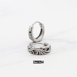 May include: Two silver-colored hoop earrings with an engraved floral design. The earrings are displayed on a white surface. The brand name "Black Fox" is printed in a black rectangle.