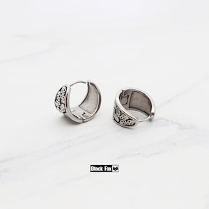 May include: A pair of silver-toned hoop earrings. Each earring features a skull pattern on the outer surface. The earrings are displayed on a white marble surface. The brand name "Black Fox" is printed in a black rectangle.