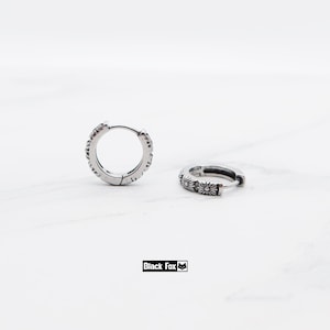 May include: Two silver hoop earrings with a floral design. The earrings are made of a shiny metal and have a detailed pattern around the edges. The earrings are shown on a white marble surface.