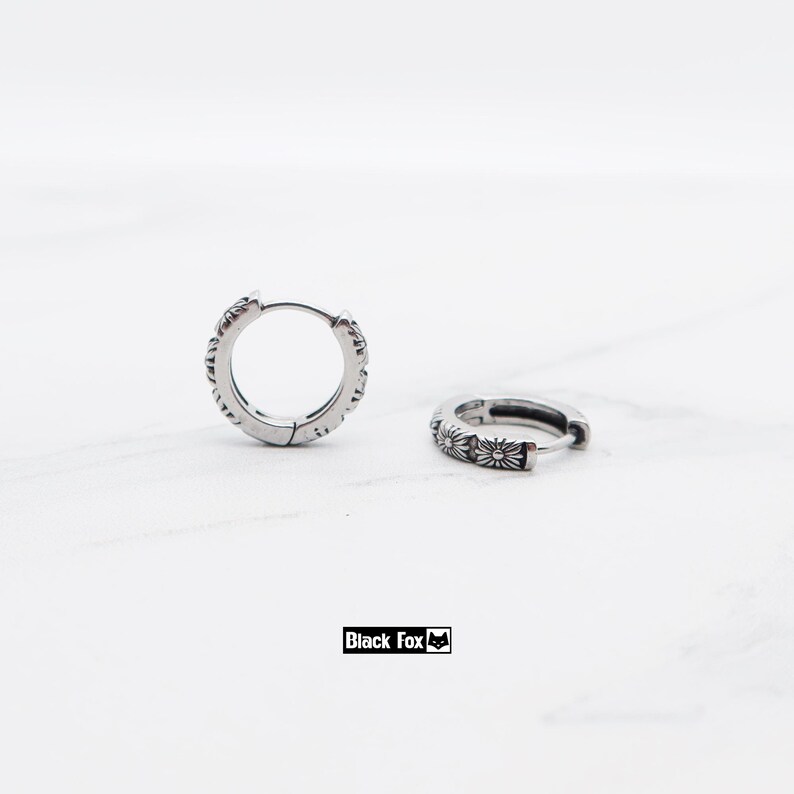 May include: Two silver hoop earrings with a floral design. The earrings are made of a shiny metal and have a detailed pattern around the edges. The earrings are shown on a white marble surface.