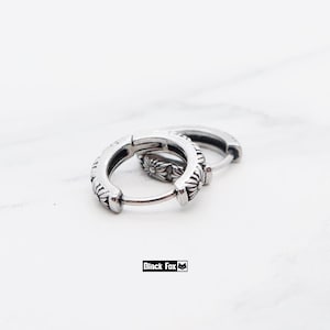 May include: Two silver hoop earrings with a floral pattern. The earrings are made of metal and have a hinged clasp. The brand name "Black Fox" is printed in a black rectangle.