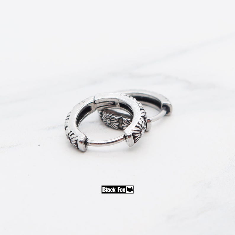 May include: Two silver hoop earrings with a floral pattern. The earrings are made of metal and have a hinged clasp. The brand name "Black Fox" is printed in a black rectangle.
