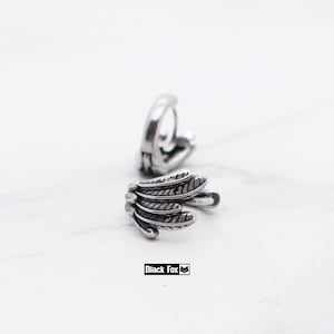 May include: Two silver feather-shaped earrings. The earrings have etched lines, giving them a textured appearance. The earrings are on a white surface. The text "Black Fox" is visible.