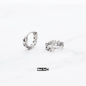 May include: Two silver hoop earrings, each adorned with a series of star-shaped cutouts. The earrings are displayed on a white surface. The brand name "Black Fox" is printed in a black rectangle.