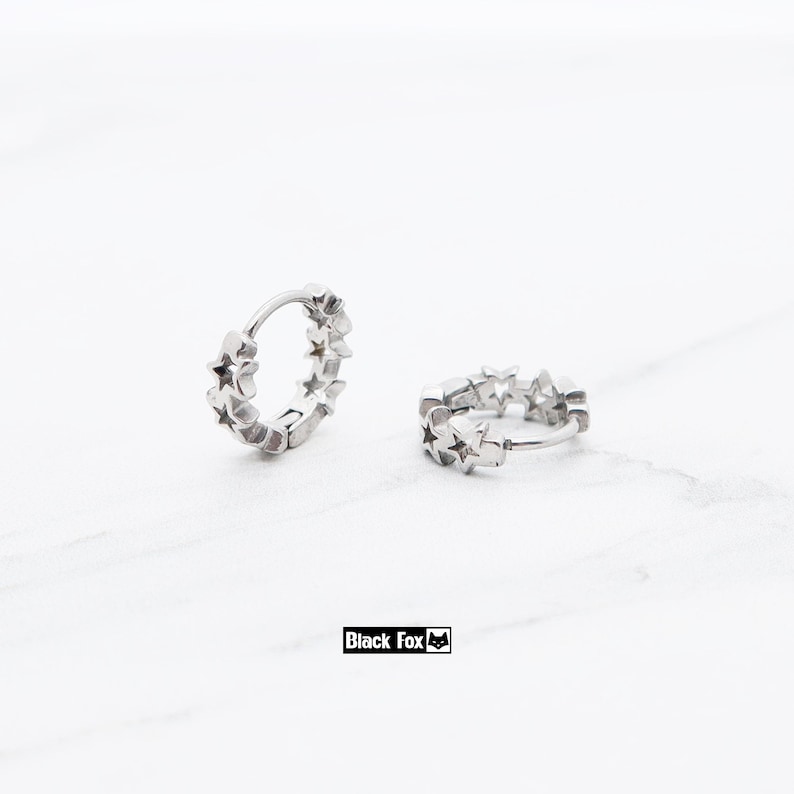 May include: Two silver hoop earrings, each adorned with a series of star-shaped cutouts. The earrings are displayed on a white surface. The brand name "Black Fox" is printed in a black rectangle.