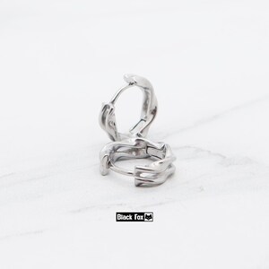 May include: A pair of silver-toned hoop earrings featuring a textured, irregular, and organic surface design. The earrings are circular with a hinged closure mechanism, showcasing a polished metallic finish that reflects light across the uneven contours.